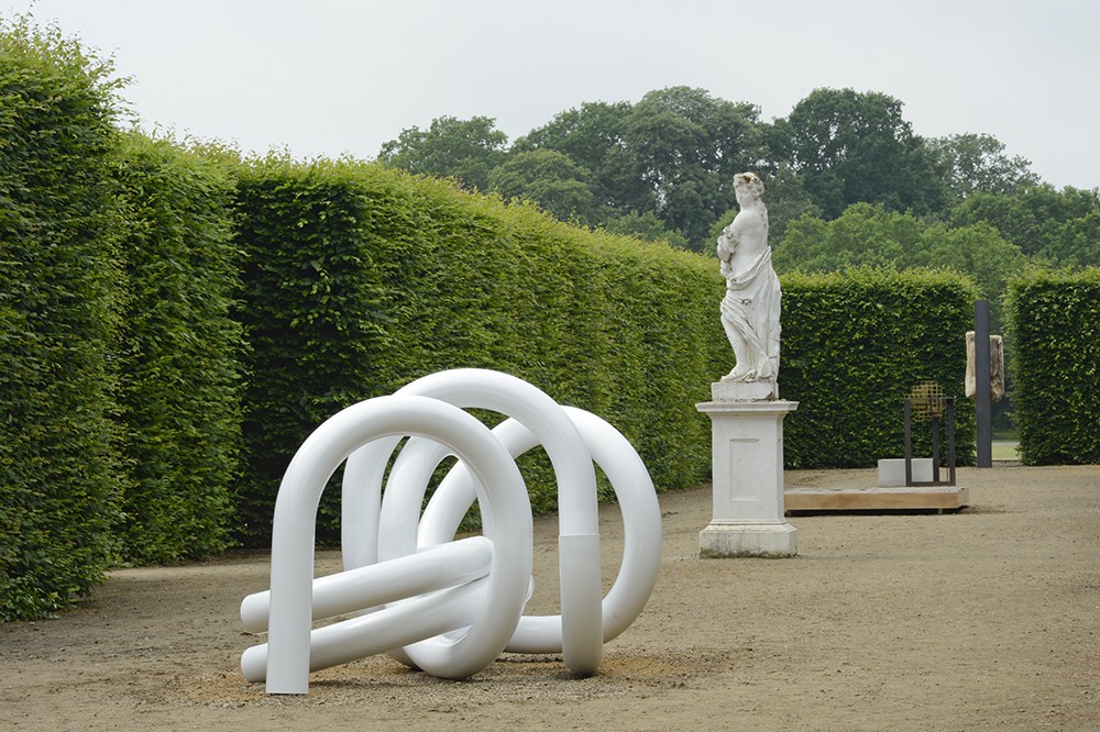 View of a curving abstract sculpture made of white-painted steel in a formal garden alongside a more classical statue