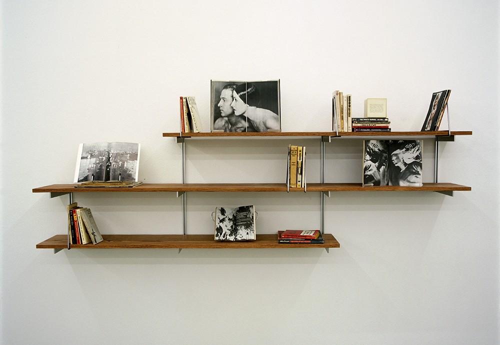 A sculpture composed of objects, including books and magazines, resting on three wooden bookshelves mounted to the wall