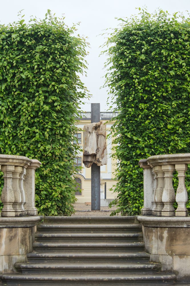 View of a sculpture made of petrified wood and a steel I-beam seen through shrubbery in a formal garden