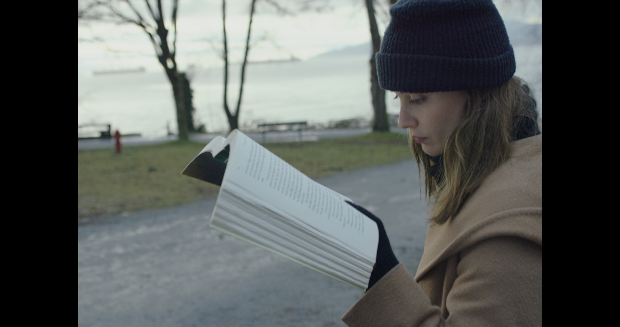 A still from Mark Lewis's 2017 film Canada depicting a young woman, in a coat, hat, and gloves, reading a book near the waterfront.