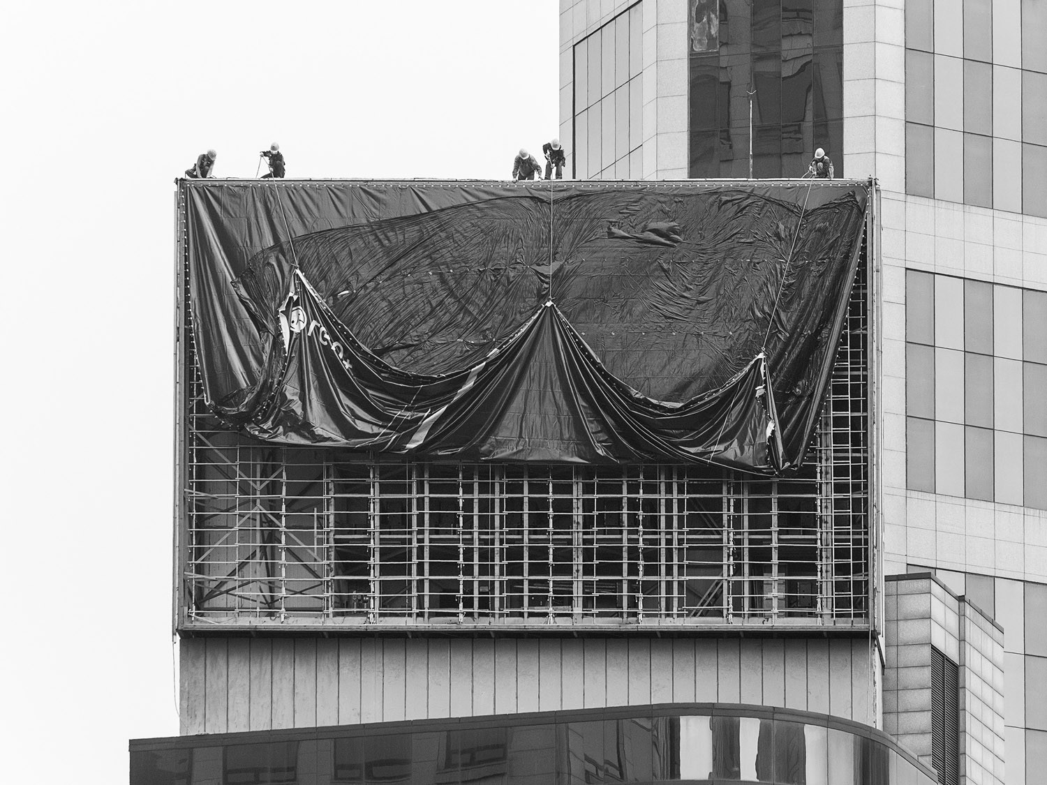 A black-and-white photograph of five men in hard hats lifting a platic tarp from the surface of a billboard