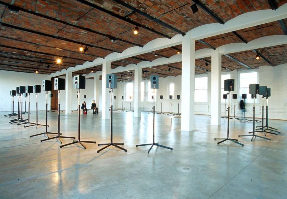 A large room with a barrel-vaulted brick ceiling contains forty music speakers on individual stands arranged in a large circle.